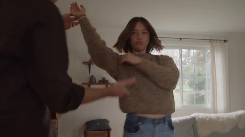 A woman in a brown sweater twirls joyfully in a cozy living room as a partner dances with her