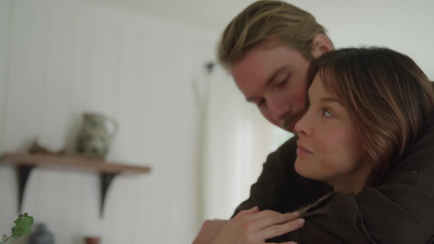 A woman in a chunky knit sweater adjusts her hair in soft indoor light as a male comes up and hugs her gently