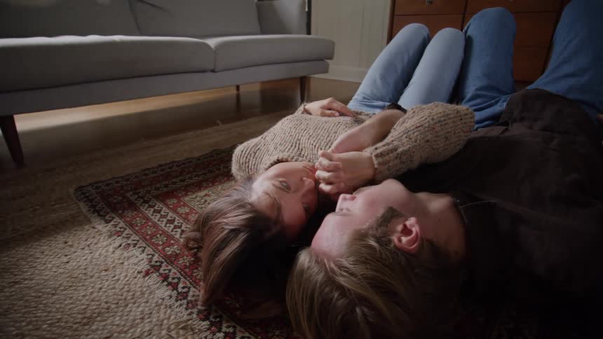 A man and a woman lie horizontally on their backs on a small patterned rug over a woven carpet in a living room looking at each other with the woman briefly placing her hand over the mans mouth and a sofa and wood furniture visible in the background