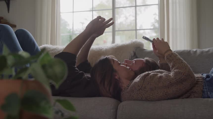 A man and a woman lie together on a cozy sofa looking at each other while simultaneously holding up their mobile phones to use social media or watch videos in a brightly lit living room