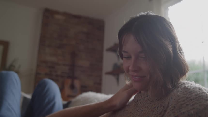 A man lies with his head resting in a womans lap on a sofa near a window while the woman smiles and gently touches his hair and face with her hand as her head is illuminated by the light from the window