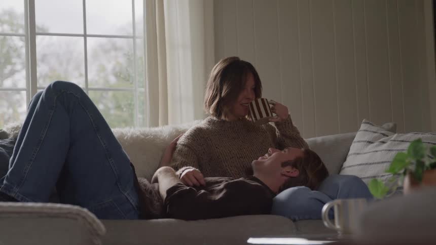 A man lies with his head resting in a womans lap on a sofa near a window looking up at the woman while she smiles down at him and drinks something from a mug