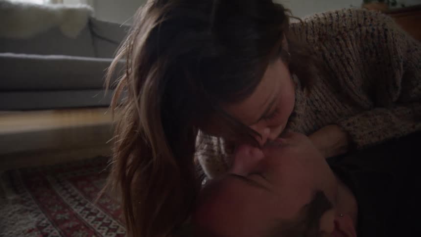 A man lies on his back on a patterned floor while a woman leans over him and both are kissing in an indoor room with unfocused furniture visible in the background