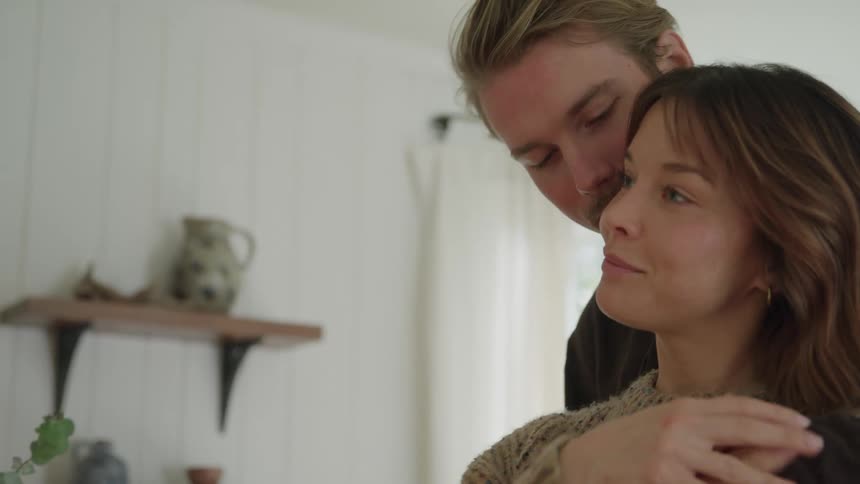 A close intimate view shows a woman in a brown sweater tucking her hair behind her ears before a man in a dark shirt approaches from behind and embraces her with a tender kiss on the cheek