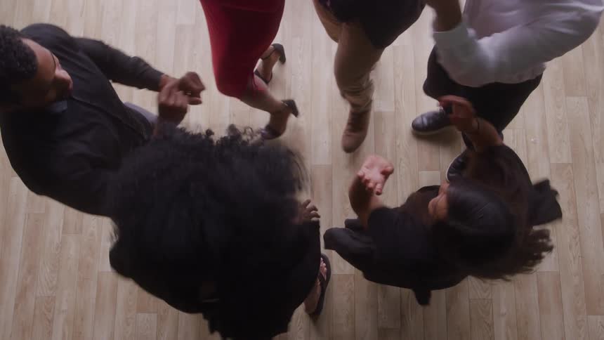 Four men and two women are viewed from directly above as they stand close together on a lightcolored wood floor and move their bodies and arms in a circular arrangement