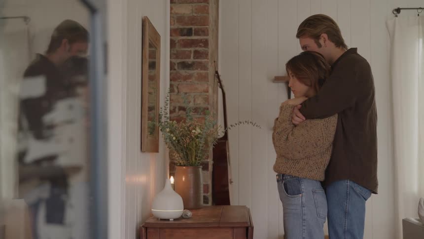 A medium shot shows a man walking up behind a woman in a brown sweater and jeans to embrace her from behind while standing near a small table with a vase of dried flowers and an oil diffuser