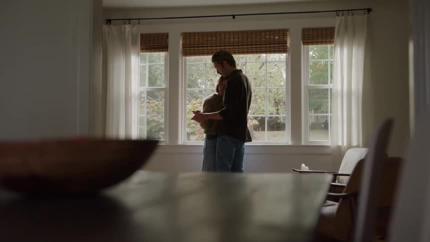 A man and a woman are in a slow dance by a large window with white curtains and bamboo blinds facing the light and embracing while dark furniture is visible in the unfocused foreground