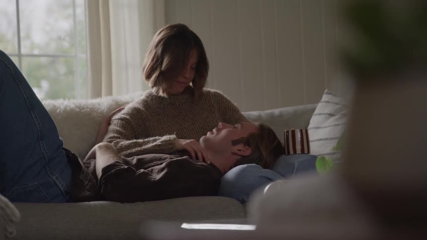 A woman sits on a couch cradling a mans head in her lap while they share a quiet intimate moment