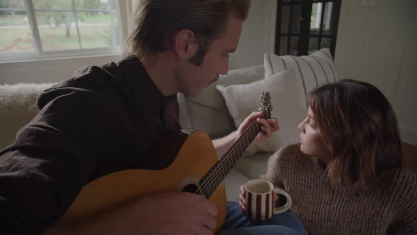 A medium view of a man playing an acoustic guitar indoors for a woman who smiles and holds a striped mug while sitting on the floor by a cozy sofa with pillows and a large window overlooking the outdoors