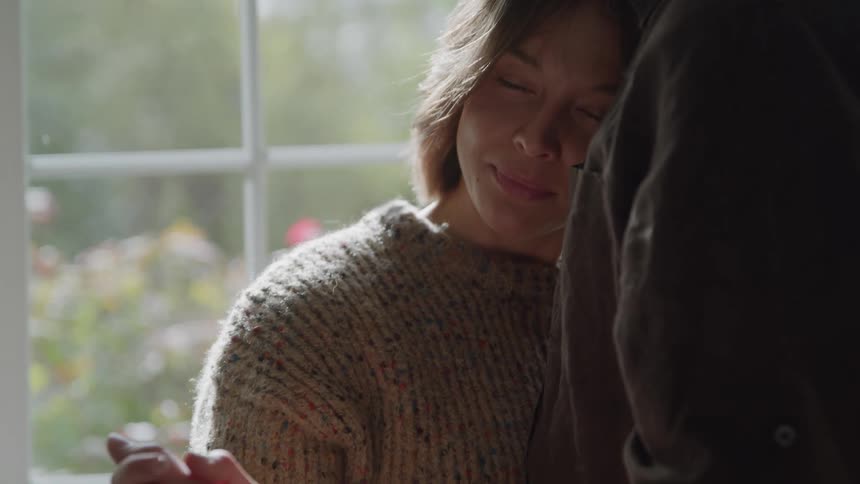A man and a woman stand close to each other in front of a window with the woman resting her head and eyes closed against the mans shoulder as they interlock their fingers and hold hands together