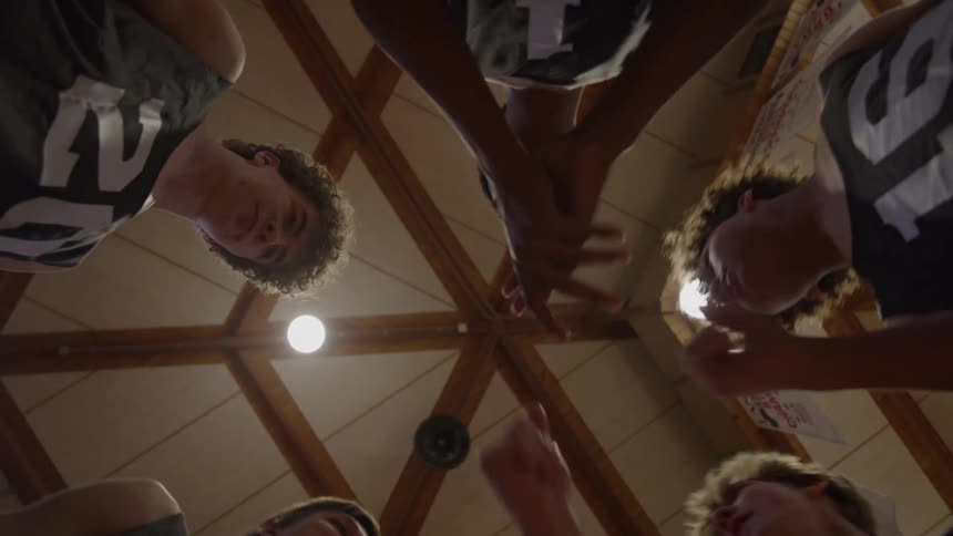 A circle of athletes huddles with hands stacked in the center as they look down with energized determination beneath wooden rafters