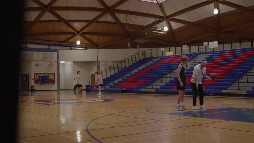 An older man and a younger man stand on a brightly lit wood gymnasium floor with the older man holding a basketball and demonstrating a movement to the younger man while other players move in the distance