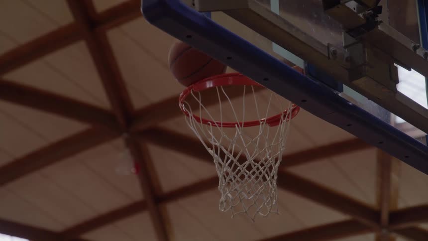 A brown basketball is thrown upward and passes through the orange hoop and white net which hangs from a blue support structure all framed against the high wooden ceiling of a gymnasium