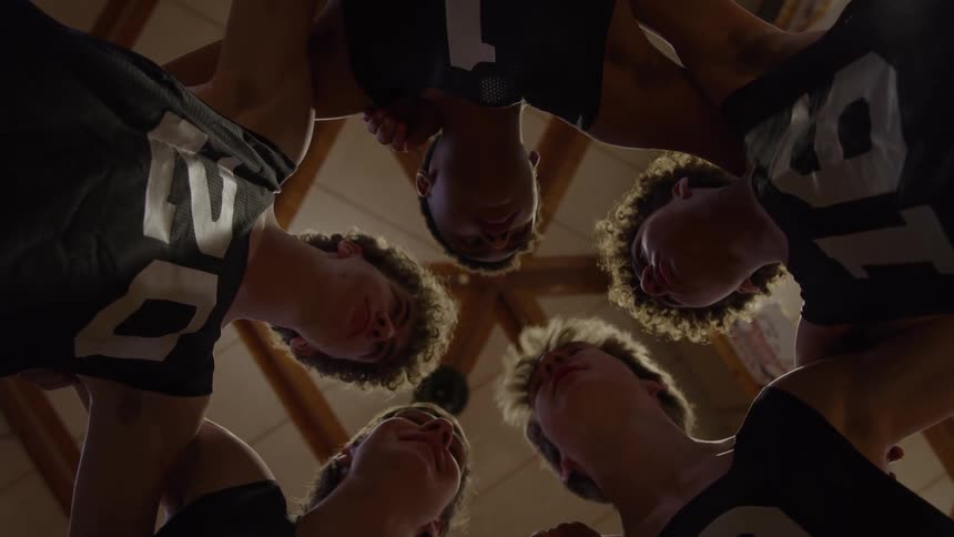 A sports team teenage boys wearing dark numbered jerseys lean their heads inward to form a circle under a high wooden ceiling with exposed beams and recessed lights