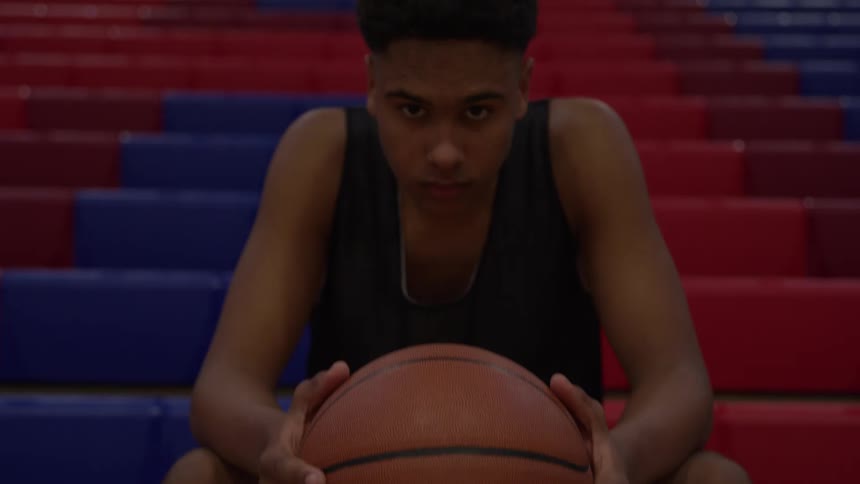 A young man wearing a dark sleeveless jersey sits facing the camera on a section of tiered seating holding a basketball against a background of red and blue bleachers