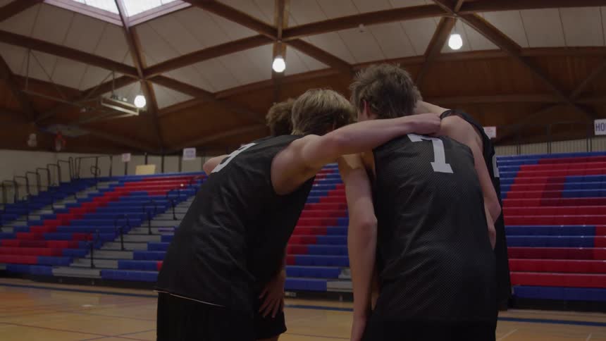 Teenage boys wearing dark numbered jerseys walk to the center of a wooden court then quickly gather in a tight circle with their arms around one another before breaking apart all in front of wide rows of empty gymnasium seating