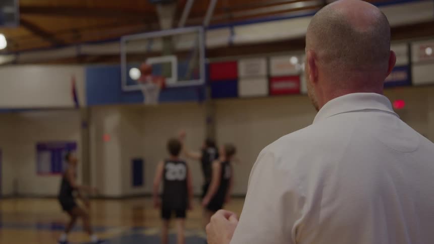 A view from over the shoulder of a bald coach in a white shirt shows him clapping and watching three young male players in black jerseys playing basketball on the court with a hoop in the background