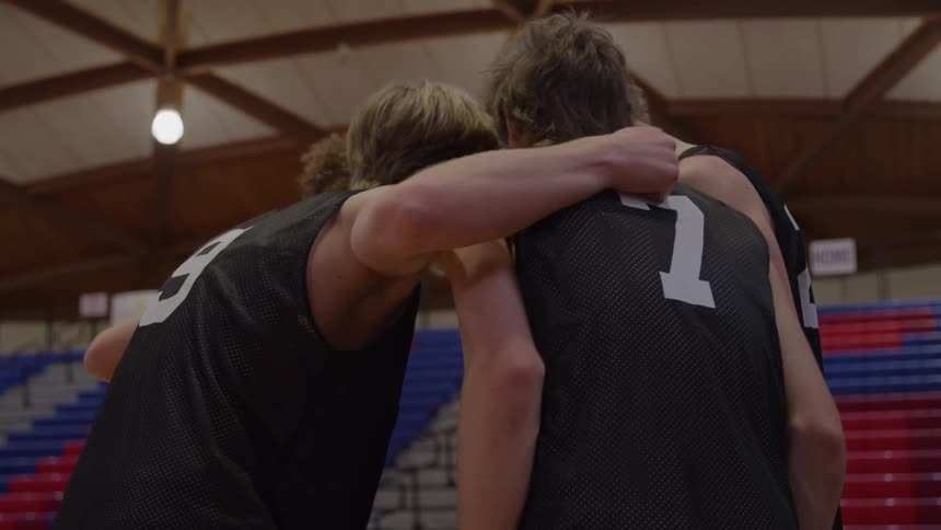 A medium view of four young male basketball players in black jerseys briefly huddling together on the court for encouragement before breaking away to play with the bleachers visible in the background