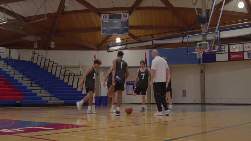 A basketball coach in a white shirt leads four young male players in black jerseys through stretching exercises on a wooden gymnasium court with blue and red bleachers in the background