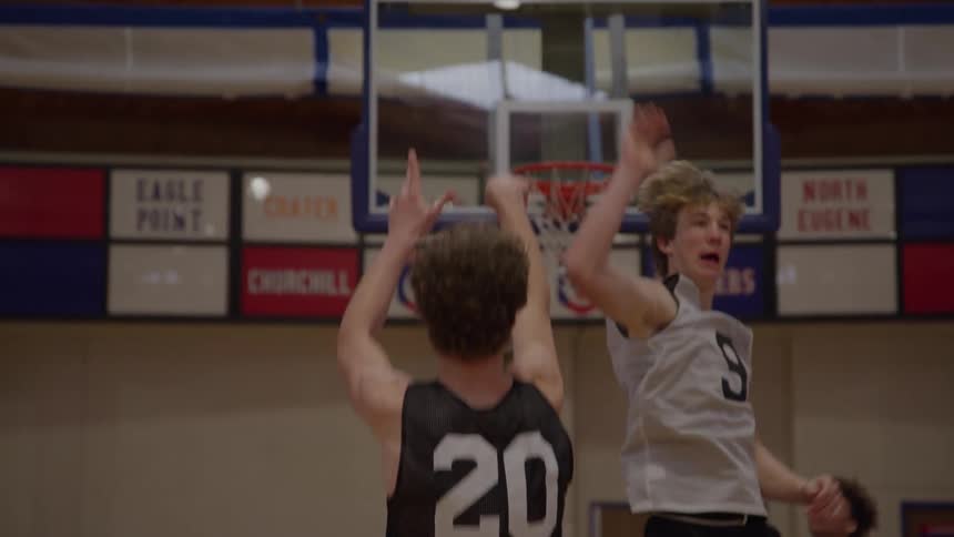 A basketball player wearing a dark jersey with the number 20 shoots an orange basketball that travels through the hoop and net while other players stand nearby on a wooden court floor in a gymnasium