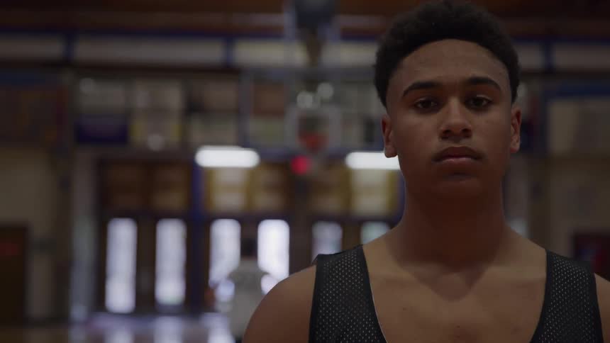 A serious young man wearing a black basketball jersey stands still in a gymnasium looking directly at the camera with a basketball hoop visible in the softly lit background