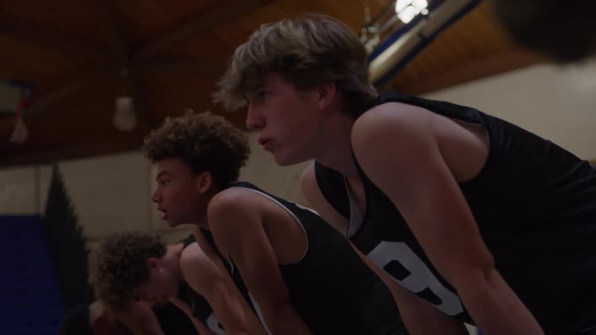 A medium side profile view of four young male basketball players in black jerseys crouching low and concentrating on a drill in a dimly lit gymnasium