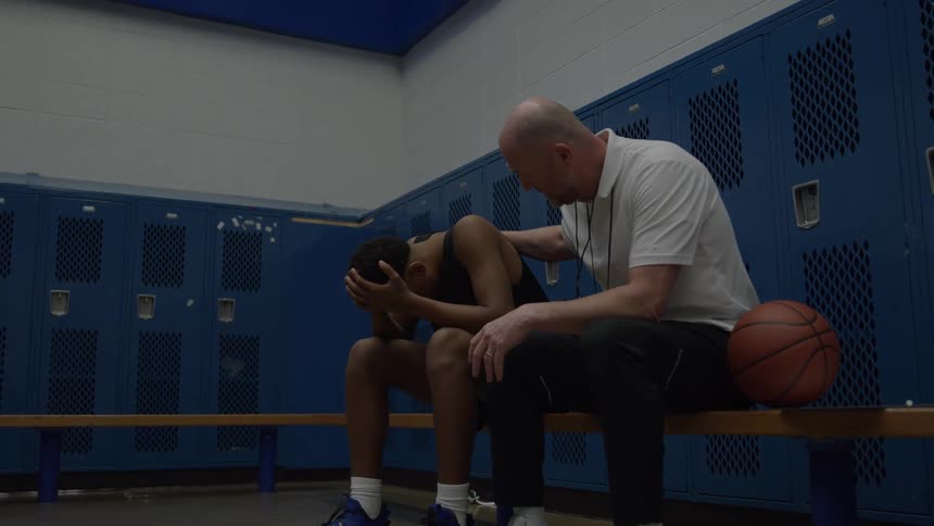 A younger man sits hunched over with his face covered by his hands on a wooden bench in a room lined with rows of closed blue metal storage compartments while an older bald man sits next to him with his hand on the younger mans back