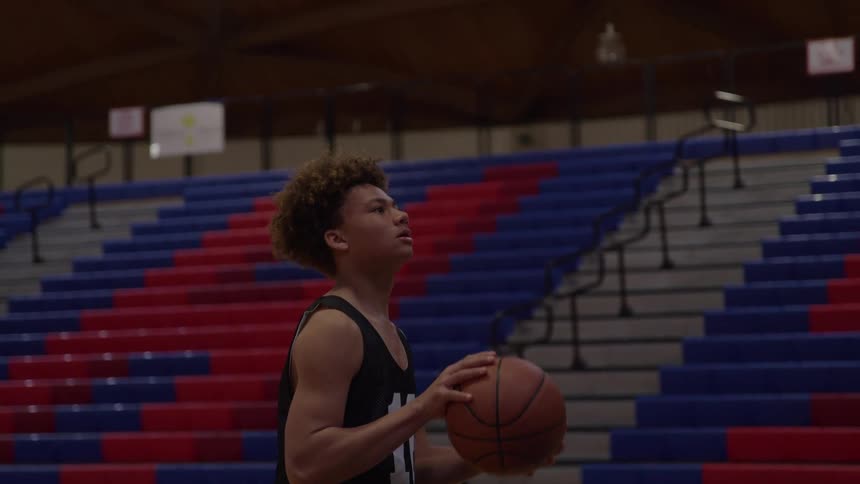 A male youth wearing a black tank top holds a brown basketball and moves the ball upward to shoot while standing on a gymnasium floor in front of rows of red and blue tiered seating