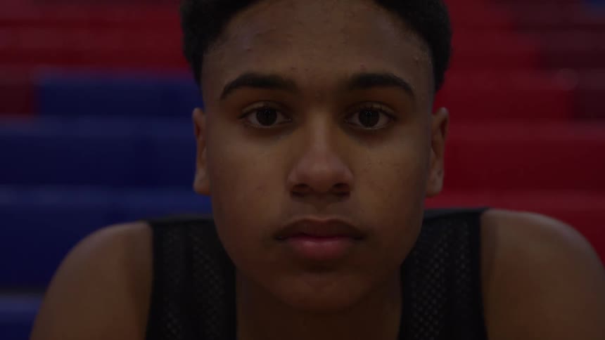 A very close view of a young man with dark hair and a black tank top slowly looks up staring intently at the camera against a blurred background of red and blue bleachers