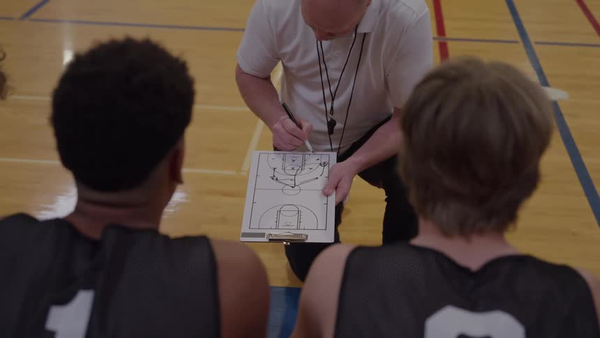 A medium rear view over the shoulders of two young basketball players in black jerseys shows a bald coach in a white shirt kneeling to draw and explain a play on a clipboard showing a basketball court diagram