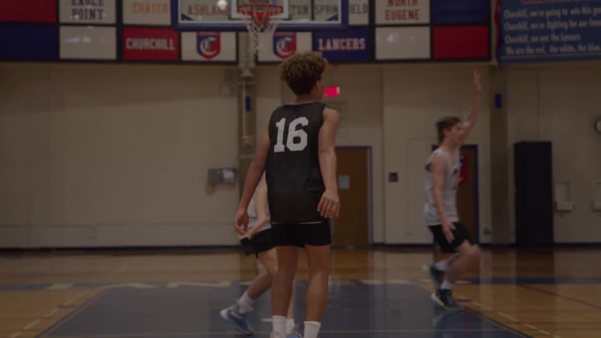 A basketball player wearing a dark jersey with the number 16 moves on a wooden court floor while holding and passing an orange ball near a defending player with a basketball hoop centered above them