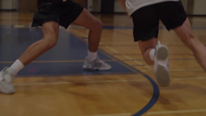 A basketball player dribbles a basketball then moves quickly past a defender and successfully throws the ball through the orange hoop and net in a gymnasium setting