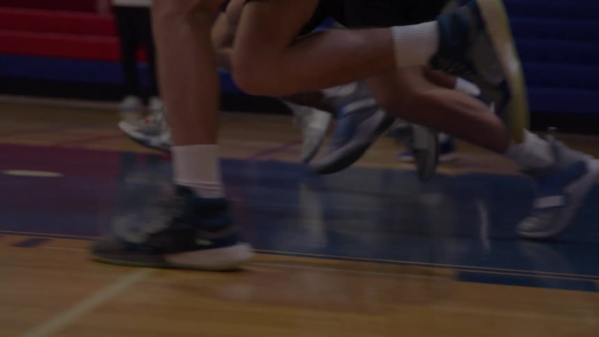 The lower legs and feet of several individuals wearing athletic shoes and white socks run rapidly across a glossy wooden gymnasium floor marked with red and blue lines with a section of red and blue tiered seating visible in the background