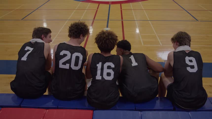 A low angle rear view shows five young male basketball players sitting on the side of a wooden court with their backs to the camera wearing black jerseys with numbers seven twenty sixteen one and nine