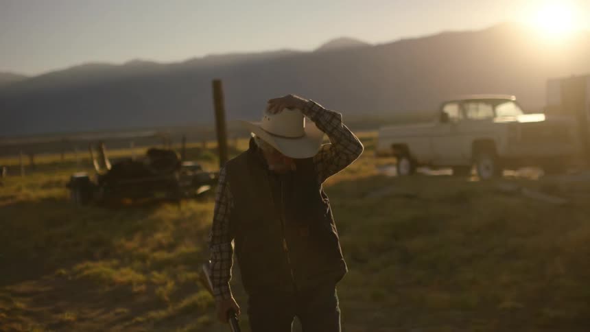 An old man is carrying a rifle as he walks outside at sunset