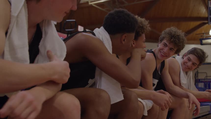 A group of four smiling young male basketball players wearing black jerseys and white towels around their necks sit close together on a bench laughing and talking in a dimly lit gymnasium
