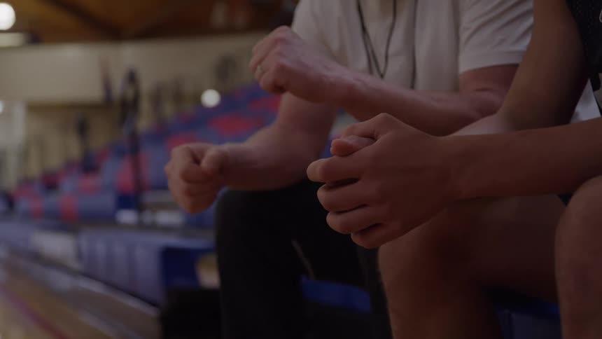 A closeup shot focuses on the hands and laps of two individuals one wearing a white shirt and the other a dark jersey as they discuss and greet each other while sitting on a bench in front of rows of red and blue tiered seating