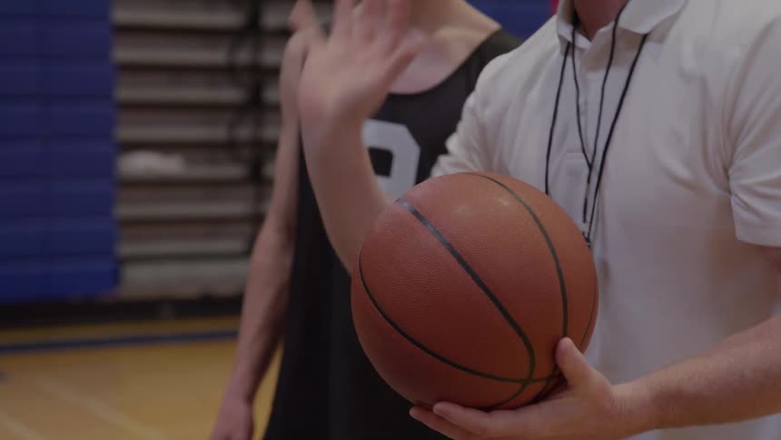 A medium close view of a bald coach in a white shirt demonstrating a move with a basketball to a young male player in a black number nine jersey on a wooden gymnasium floor