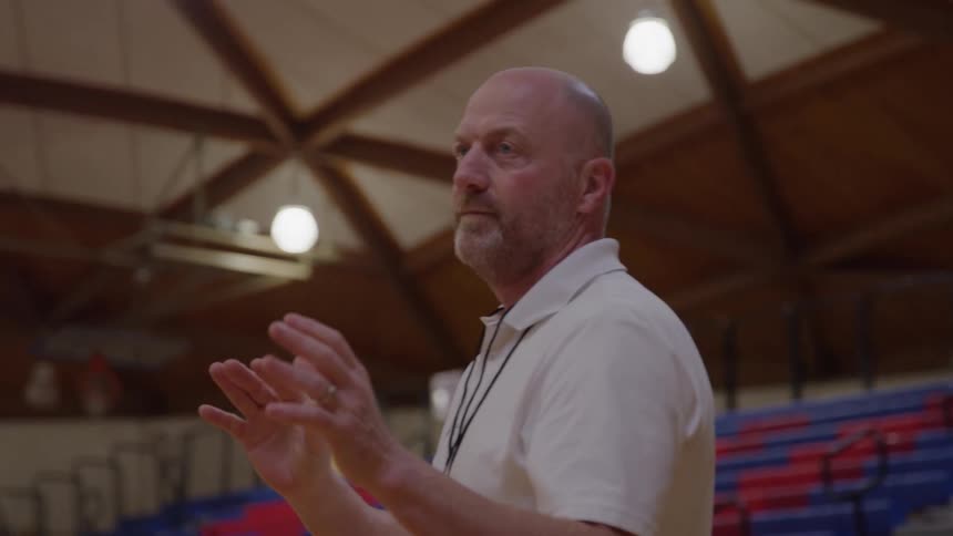 A bald man wearing a white collared shirt stands on a gymnasium court in front of tiered red and blue seating clapping his hands together and gesturing while speaking then catches an orange basketball thrown toward him
