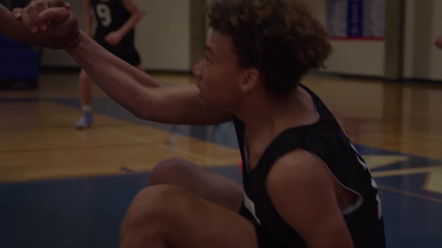 A young male basketball player in a black number sixteen jersey sits on the court before accepting a hand from a teammate to stand up and join the fast paced game