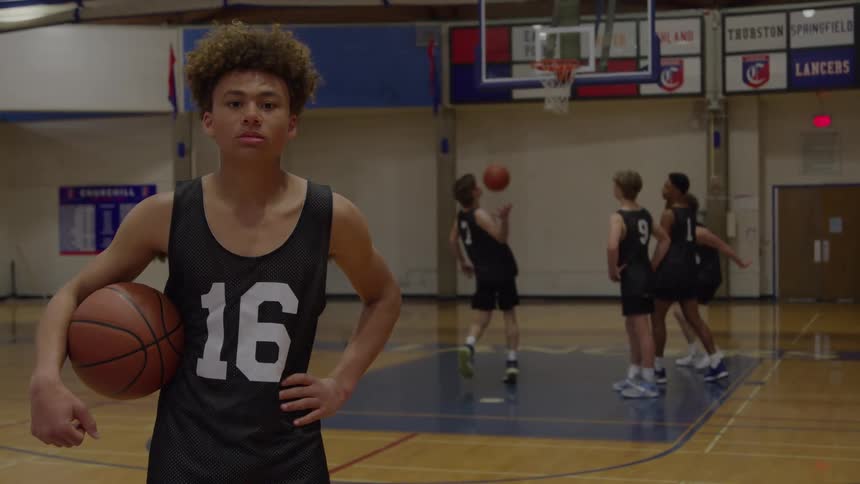 A male teen athlete posing with a ball in front of playing teammates in a basketball court