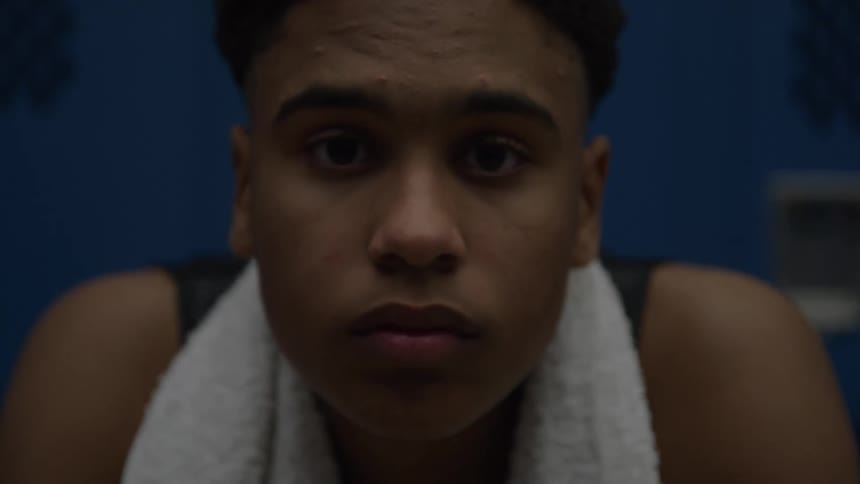A young man with a towel around his neck looks straight ahead in a locker room with a sad slightly pensive expression