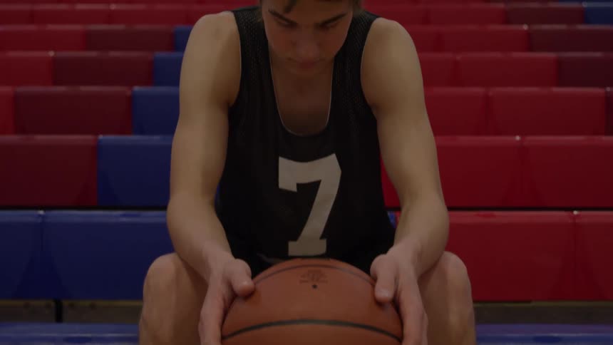 A young male basketball player wearing a dark numbered jersey sits on a section of red and blue tiered seating holding a basketball and slowly lifts his head to look directly at the camera