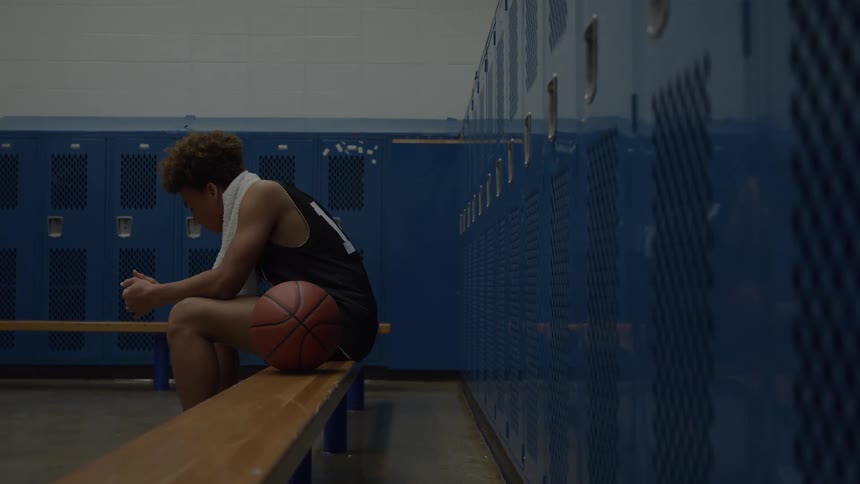 A basketball player with a towel around his neck sits hunched on a locker room bench with the ball at his side looking exhausted and pensive