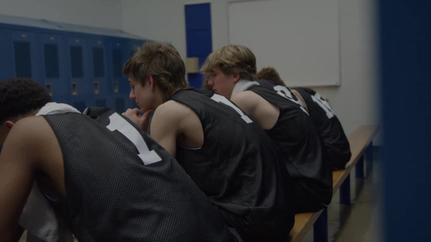 A side view of four young male basketball players in black jerseys sitting quietly on a wooden bench looking down with expressions of disappointment in a dimly lit blue locker room