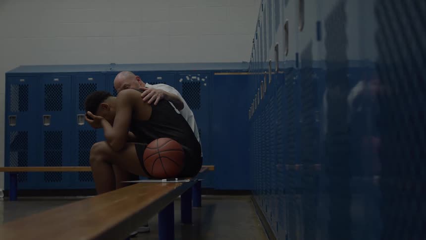 A younger man sits hunched over with his head in his hands on a long wooden bench in a room lined with blue metal storage compartments while an older bald man stands behind him and places a hand on his back as a basketball rests next to him