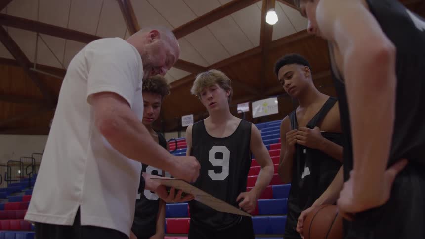A medium view of a bald coach in a white shirt pointing at a clipboard while giving instructions to four young male basketball players in black jerseys who stand focused in a gymnasium