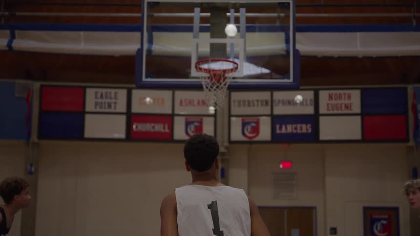 A basketball player wearing a white jersey with the number 1 stands with his back to the camera on an indoor court bounces a brown ball then raises the ball to shoot it toward the basketball hoop with other people playing
