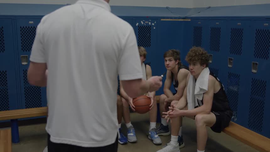 A basketball team sits on the bench in the locker room while their coach speaks