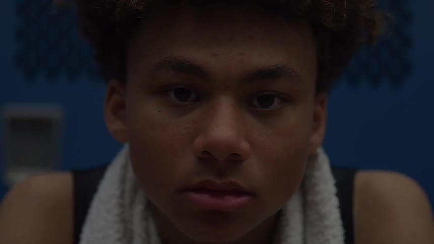 A serious young man in a black tank top with curly brown hair and a white towel draped around his neck slowly looks up and stares intently into the camera in a darkly lit locker room or gym